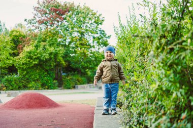 Mavi şapkalı neşeli küçük bir çocuk, sonbahar renkleriyle dolu canlı bir parkta yemyeşil çalılarla çevriliyken neşe içinde dengede duruyor..