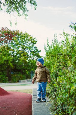 Neşeli genç bir çocuk yemyeşil yemyeşil ile çevrili canlı bir parkta alçak bir duvarda duruyor. Açık havada eğleniyor, aydınlık bir gün için sıcak giyinmiş..