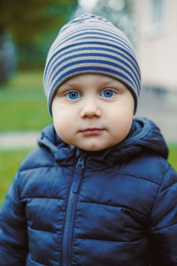 Çarpıcı mavi gözlü çocuk serin bir günde dışarıda duran sıcak mavi bir ceket ve çizgili bir şapka giyer. Yemyeşil arka plan, sahneye ferahlatıcı bir dokunuş ekler..