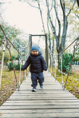 Genç bir çocuk sakin bir parktaki sağlam ahşap bir köprüde güvenle yürür. Sonbahar yaprakları, serin hava çevreyi doldururken, sakin manzaraya altın bir dokunuş ekler..