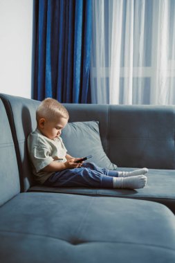 Küçük bir çocuk, yumuşak mavi bir kanepede rahat bir şekilde oturur ve akıllı bir telefona odaklanır. Güneş ışığı perdelerin arasından akar, rahat yaşam alanını doldurur..