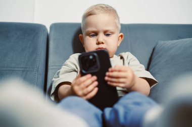 Meraklı bir çocuk, lüks mavi bir kanepede rahat bir şekilde oturan akıllı bir telefona odaklanmış. Güneş ışığı pencereden akar ve oyun ve öğrenme için sıcak bir atmosfer yaratır..