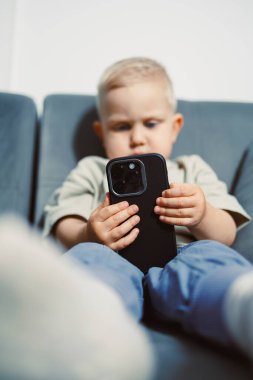 Genç bir çocuk akıllı telefonuna odaklanmış, yumuşak bir kanepede oturuyor. Açık renk saçları ve günlük kıyafetleri güneşli bir odada rahat bir öğleden sonra geçirdiği izlenimini veriyor. Ortam sakin ve davetkar..