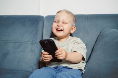 Genç bir çocuk akıllı telefonuna yaygın bir şekilde gülümsüyor, açıkça gördüklerinden memnun. O, parlak ışık ve neşeli bir atmosfer ile çevrili yumuşak bir kanepeye rahatça oturur..