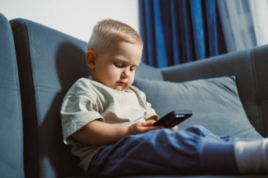 Küçük çocuk, yumuşak yastıklarla çevrili akıllı telefonuyla bir kanepede huzur içinde oturuyor. Altın öğleden sonra ışığı yakındaki pencereden içeri akıyor..