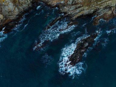 Dalgalar barışçıl bir sahil şeridindeki kayalık oluşumlara karşı şiddetli bir şekilde dalgalanır ve kırılır. Derin mavi deniz alacakaranlıkta kaybolan ışığı yansıtır. Huzurlu ama dinamik bir sahne yaratır..