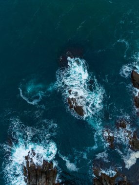 Parlak bir günbatımı altında, okyanus dalgaları engebeli kayalara çarpar ve havaya su püskürtür. Denizin canlı mavileri kayalık kıyı şeridiyle çok güzel tezat oluşturuyor..