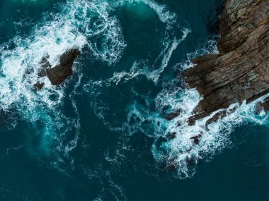 Okyanus dalgaları sivri kayalıklara karşı enerjik bir şekilde yuvarlanarak turkuaz suyun üzerinde dans eden beyaz köpük oluşturur. Güneş ışığı yüzeyden yansıyarak canlı renkleri arttırır..