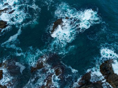 Güneş mavi sularda dans ederken köpüklü dalgalar karanlık, pürüzlü kayalar üzerinde yuvarlanır. Bu sakin sahil manzarası doğanın gücünü ve sakinliğini yansıtıyor..