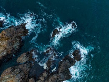 Dalgalar, dramatik bir kıyı şeridi boyunca uzanan pürüzlü kayalara şiddetle çarparlar. Gün geceye geçerken güneş ışığı, sahnenin üzerine sıcak bir parıltı saçar..