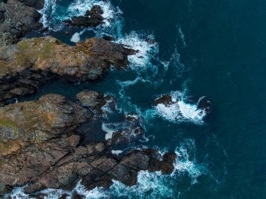 Dalgalar enerji ve güzellikle dolu bir kıyı sahnesinde karanlık kayaların üzerinde yuvarlanır. Derin mavi deniz engebeli kıyı şeridiyle kesişiyor ve doğanın gücünü hayret verici bir şekilde gösteriyor..
