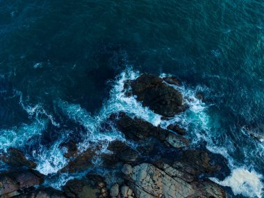 Parlak mavi dalgalar, kıyı boyunca uzanan sivri kayalara çarparak öğleden sonra erken saatlerde sıcak güneş ışığında köpürerek doğanın saf güzelliğini ve gücünü gözler önüne serer..