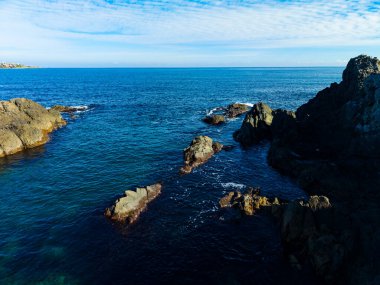 Dalgalar kayalık kıyı şeridinde yavaşça dolaşarak berrak suya güneş ışığını yansıtır. Parlak mavi bir gökyüzü ve ince bulutların üzerinde süzüldüğü gün huzur dolu..
