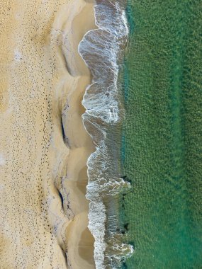 Güzel bir plaj sahnesi, altın kumu yalayan yumuşak dalgalarla gözler önüne seriliyor. İzler, sakin denizin gün batımında ufukla buluştuğu parlak kıyı şeridine doğru yol alıyor..