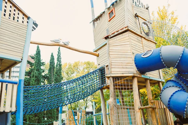 Çocuklar, ağaçlar ve sonbahar yapraklarıyla çevrili canlı bir şehir parkında yaratıcı ahşap yapılarda tırmanıp oynarken ve renkli bir kaydırak üzerinde görülebilir..