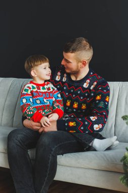 Neşeli bir baba ve oğul, hem renkli tatil kazaklarıyla kanepede, bu şenlikli sezonda birlikteliğin ruhunu kutlayarak, yürek ısıtan bir anı paylaşırlar.