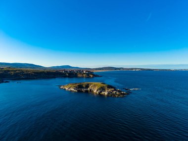 Dalgalar, parlak gökyüzü altında karası olan küçük kayalık bir adanın etrafında yavaşça hareket eder. Uzak tepeler bulutsuz güneşli bir günde arka planda..