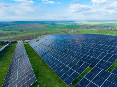 Büyük güneş paneli kurulumu açık bir gökyüzünün altındaki yeşil arazide bulunuyor. Uzak tepeler görünürken güneş panellerin üzerinde parlıyor. Bu alan yenilenebilir enerji kullanımını gösterir.