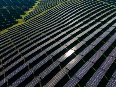 Güneş panelleri geniş yeşil bir alanda desenler halinde dizilmiştir. Parlak güneş panellerde parlıyor, yenilenebilir enerji üretimi üzerine odaklanmış bir sahne yaratıyor..