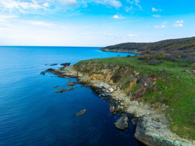 Bir kıyı şeridinde kayalık oluşumlar ve berrak mavi su bulunur. Güneş manzaranın üzerinde ışıl ışıl parlıyor, arkadaki tepeler mavi bir gökyüzünün altında görülebiliyor..