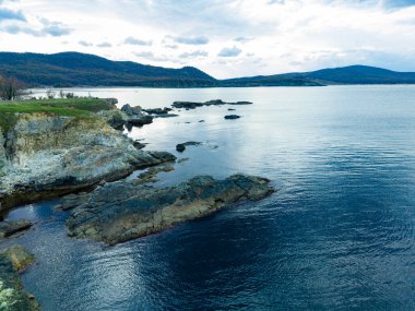 Kayalık bir kıyı şeridi, kayalara çarpan yumuşak dalgalarla suyla birleşir. Tepeler bulutlarla dolu bir gökyüzünün altında yükselir..