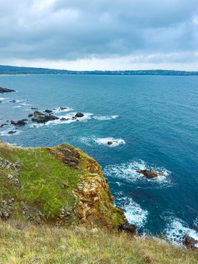 Okyanus kıyısındaki kayalıklarda dalgalar kırılıyor. Su mavi ve gökyüzünde bulutlar var. Uzaktan bir kıyı kasabası görünüyor..