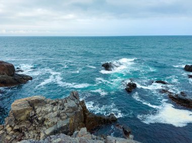 Dalgalar bulutlu bir öğleden sonra sahildeki kayalara çarpar. Su mavidir ve ufuk uzaklıktan görülebilir. Sahne doğal bir manzara gösteriyor..
