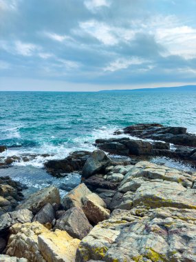 Okyanus arka planda uzanırken dalgalar kıyı boyunca kayalara çarpar. Bulutlar gökyüzünü kaplıyor ve manzara doğal bir kıyı manzarasını gösteriyor..