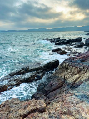 Güneş batarken dalgalar kayalık sahile vurur ve manzaralı bir manzara yaratır. Okyanus bulutlu bir gökyüzünün altında uzak dağlara doğru uzanıyor..