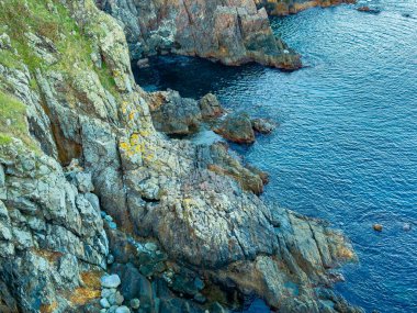 Sahne, kıyı bölgesindeki kayaları ve suyu gösteriyor. Dalgalar yavaşça taşlı kıyıya dokunur. Su berrak ve mavi. Güneş doğanın üzerine parlıyor, parlak bir atmosfer yaratıyor..
