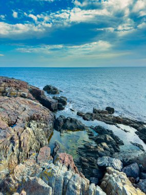 Suyun karayla buluştuğu kıyıda kayalar var. Dalgalar taşların üzerine akar. Bulutlar denizin üzerinde yüzer. Sahne doğanın güzelliğini ve suyun hareketini gösteriyor..