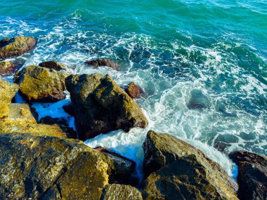 Dalgalar kayalık sahile vururken su taşlara vuruyor, güneşli bir günde okyanusun hareketini parlak renkler ve berrak görüş mesafesi ile gösteriyor..
