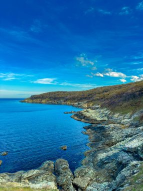 Bir kıyı şeridi su boyunca uzanır ve kayalar sarsılır. Yukarıdaki gökyüzü birkaç bulutla mavi. Arka planda yeşil tepeler yükseliyor. Öğlen oldu ve sahne aydınlık ve açık..