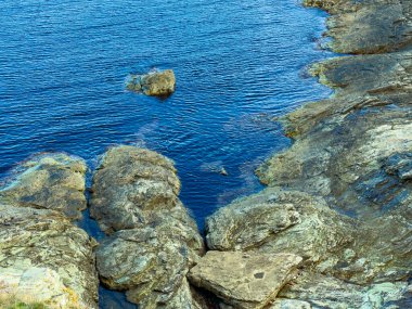 Sahne, öğleden sonra açık mavi suyu olan kayalık bir kıyı şeridini gösteriyor. Suyun üzerinde büyük taşlar görülebilir, doğal bir manzara yaratır..