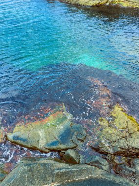 Manzara, güneş ışığını yansıtan berrak suyu ve yüzeyin hemen altındaki renkli kayaları gösteriyor. Yumuşak dalgalar kıyıya vurur ve canlı bir kıyı atmosferi yaratır..