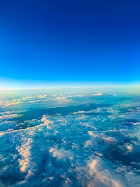 Beyaz bulutlar açık mavi gökyüzünün altındaki zemini kaplıyor. Sahne, gündüz vakti bulutları aydınlatan güneş ışığıyla birlikte havadan bir görüntü gösteriyor..