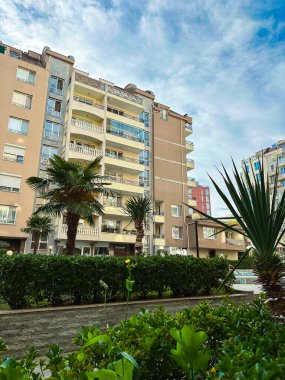Palmiye ağaçları ve çalılar gün boyunca bir apartmanın etrafını sarar. Gökyüzü parçalı bulutlu ve insanlar arka planda yürürken görülebilir..