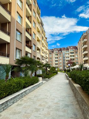 Bitkiler ve ağaçlarla kaplı bir yürüyüş yolu yerleşim alanındaki binaları birbirine bağlar. Gökyüzü açık ve mavi, üzerinde bulutlar var..