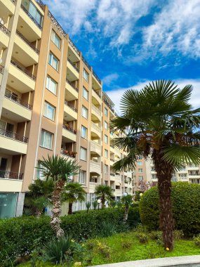 Çok katlı bir bina birçok balkona sahiptir. Palmiye ağaçları ve çalılar parlak mavi bir gökyüzünün altında binanın etrafında yeşil bir alan oluşturur..