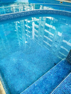 Açık mavi yüzme alanı binaların yansımalarıyla temiz su gösteriyor. Fayanslar havuzun etrafında bir desen oluşturarak gün ortasında parlak ve davetkar bir sahne yaratıyor..