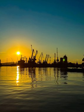 Güneş bir limanın üzerinde batar ve suyun üzerinde parlayan parlak bir ışık yaratır. Rıhtım boyunca vinç ve gemi siluetleri görülüyor..