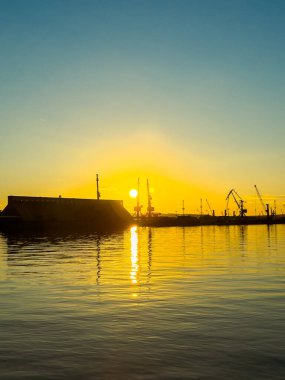 Güneş, suya altın bir parıltı saçarken limanın üzerinde batıyor. Renkli gökyüzünün arka planında vinç ve tekne siluetleri görülür..