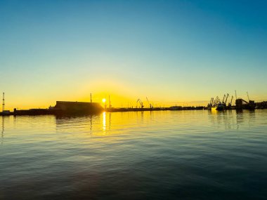 Güneş limanın arkasında batıyor, suya turuncu ışık saçıyor. Turnalar rıhtım boyunca görülebiliyor ve sahile yakın bir yerde gün boyu hareketlilik var..