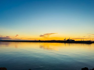 Güneş sakin suyun üzerinde batarken gökyüzü parlak renkler gösteriyor. Gece ışığında kıyı şeridi boyunca kayık siluetleri ve uzak binalar görülebilir..