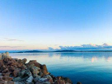 Açık bir sabah, sakin bir suyun gökyüzünü yansıttığını gösteriyor. Kıyı boyunca kayalar var ve uzak dağlar ışık bulutlarının altında görülebilir. Manzara şafak vakti doğanın güzelliğini yakalar..