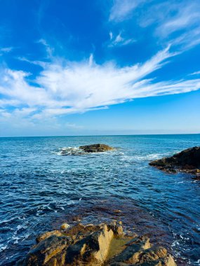 Dalgalar, üzerinde açık mavi bir gökyüzü olan kayalara çarpar. Sahne, okyanusun karayla buluştuğu güneşli bir günü yakalar..