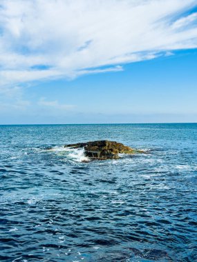 Su ve dalgalarla çevrili bir kaya okyanusta duruyor. Gökyüzü mavi renklerle parçalı bulutludur. Sahne, güneşli bir günde doğada bir anı yakalar..