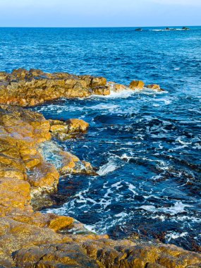 Dalgalar kıyıdaki engebeli kayalara vururken mavi okyanus berrak gökyüzünün altındaki mesafeye kadar uzanıyor. Sahne, suyun ve karanın hareketini yakalar..