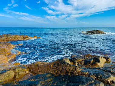 Dalgalar engebeli bir kaya kıyısına hafifçe çarpar. Güneş, çok az bulutla birlikte masmavi bir gökyüzünde parlıyor. Bu kıyı bölgesi doğanın güzelliğini ve okyanusun genişliğini gösteriyor..