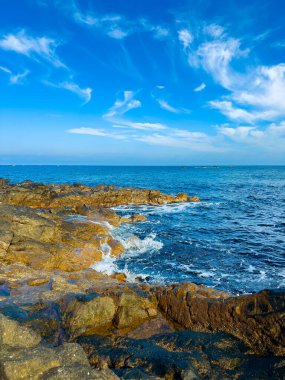 Dalgalar kıyı şeridi boyunca birkaç bulutla mavi gökyüzünün altında kayalara hafifçe çarpar. Sakin okyanus gün ışığında uzaklara doğru uzanıyor..